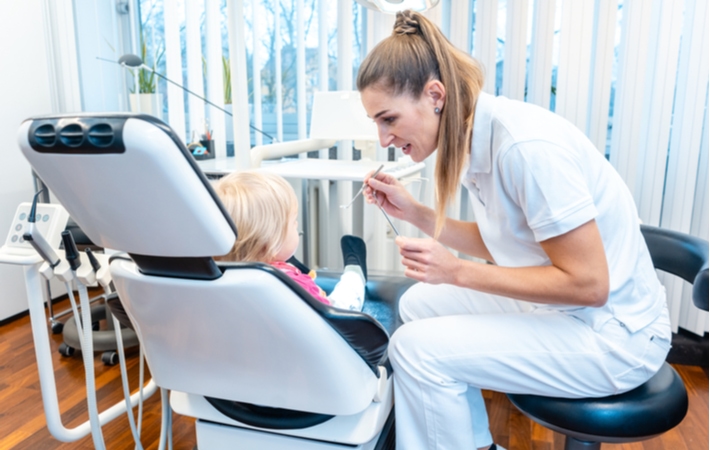 A female dentist in a white uniform preparing to examine the mouth of the small child sitting in the dental chair