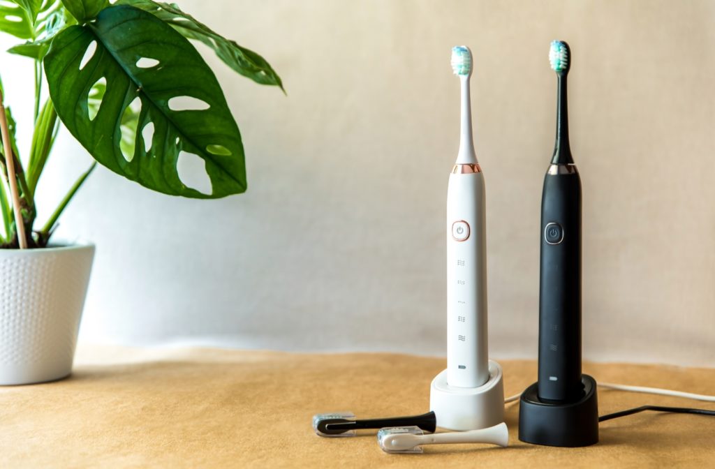 Two electric toothbrushes, one black and one white sitting on their charging stations on a wooden surface next to a potted plant