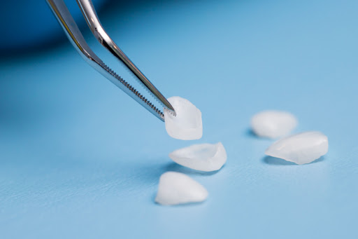 Several dental veneers laying on a blue surface with one getting picked up by tweezers.
