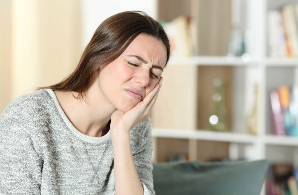 A lady in a gray shirt is sitting on a couch with a gesture of pain on her face while her left hand is touching her jaw.