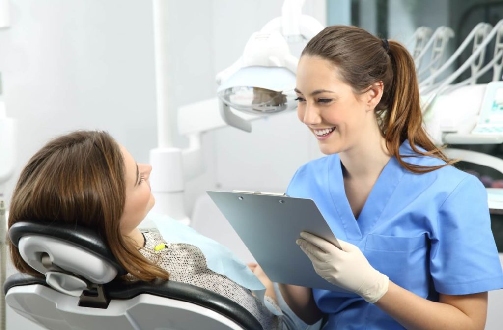A female dentist in blue scrubs and a woman sitting in a dentist's chair holding a chart and smiling.