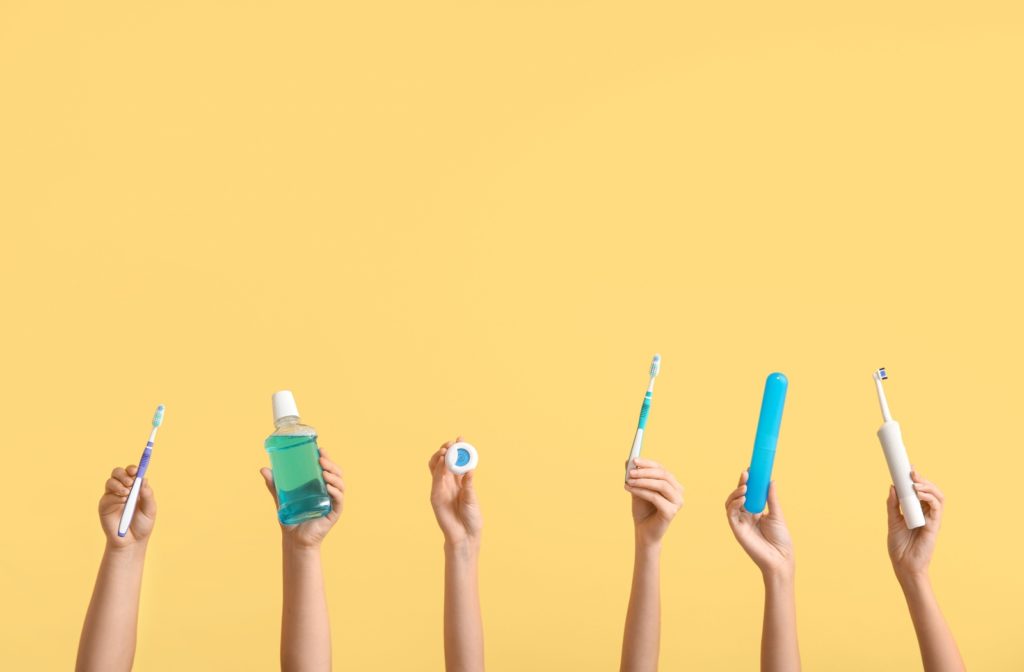 6 hands against a yellow background, each holding up a different dental care product (toothbrush, mouthwash, floss, etc.)