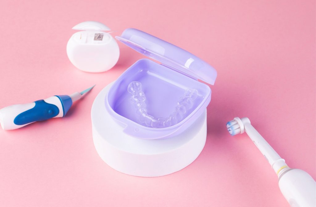 Clear aligners in a purple case resting on a small white stand with floss, an electric toothbrush, and a water flosser next to them.