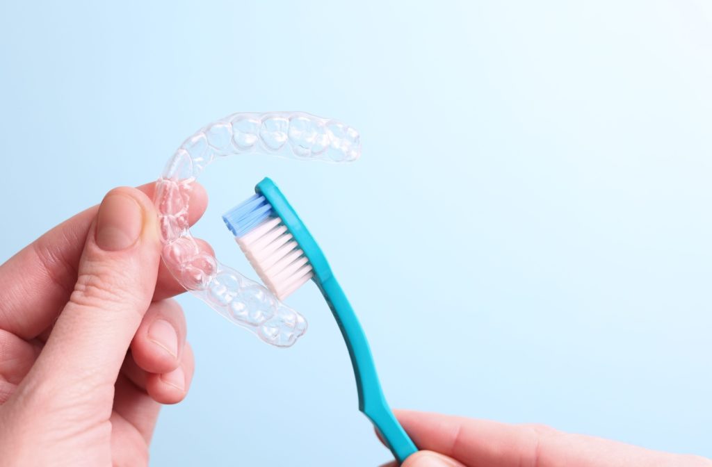 Close-up of a person's hands cleaning clear aligners with a blue toothbrush against a light blue background.