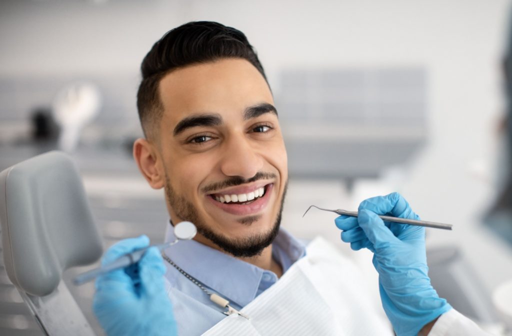 Close-up of a young man smiling brightly as he sits in a dental chair with a white bib on and a dentist holds up a small mirror and pick next to his mouth.