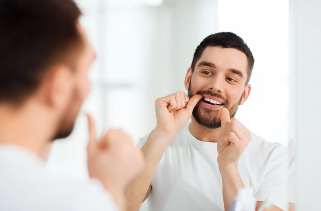 A young man wearing a white T-shirt in an all-white bathroom, flossing his teeth while looking in the mirror