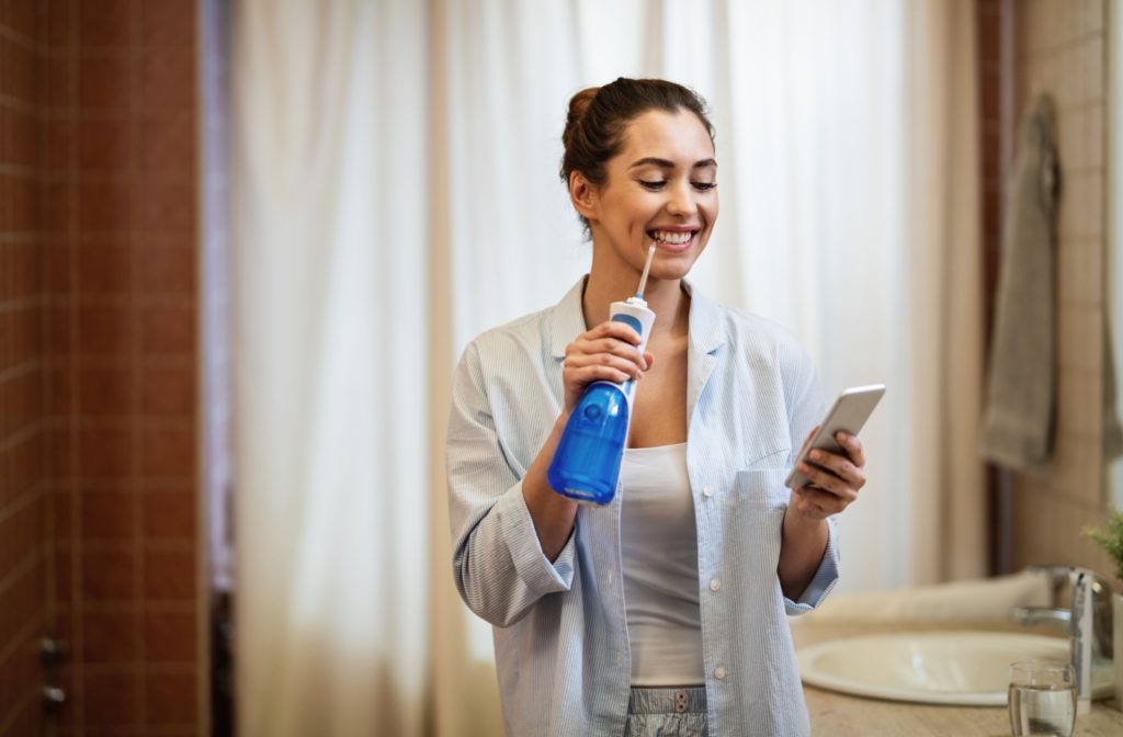 A person standing in a bathroom in pyjamas and holding their phone to set a time while using a water flosser