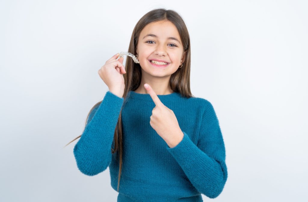 a teenager in a blue sweater holds up an Invisalign clear aligner with one hand while happily pointing to the aligner with the other hand