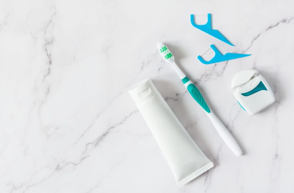  A photo of a tube of toothpaste, a toothbrush, dental floss, and two flossing sticks against a white marble background.